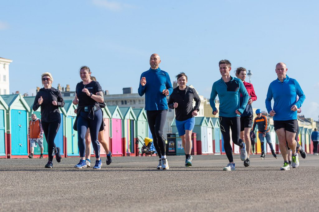 Men and women running besides a row of beach huts