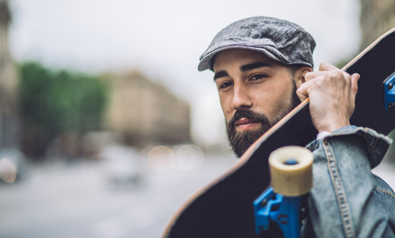 A young man with a beard holds a skateboard on his shoulder.
