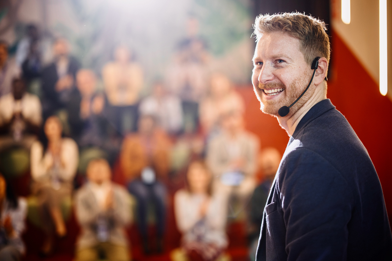 man with a headset presenting something standing in front of a large audience and smiling