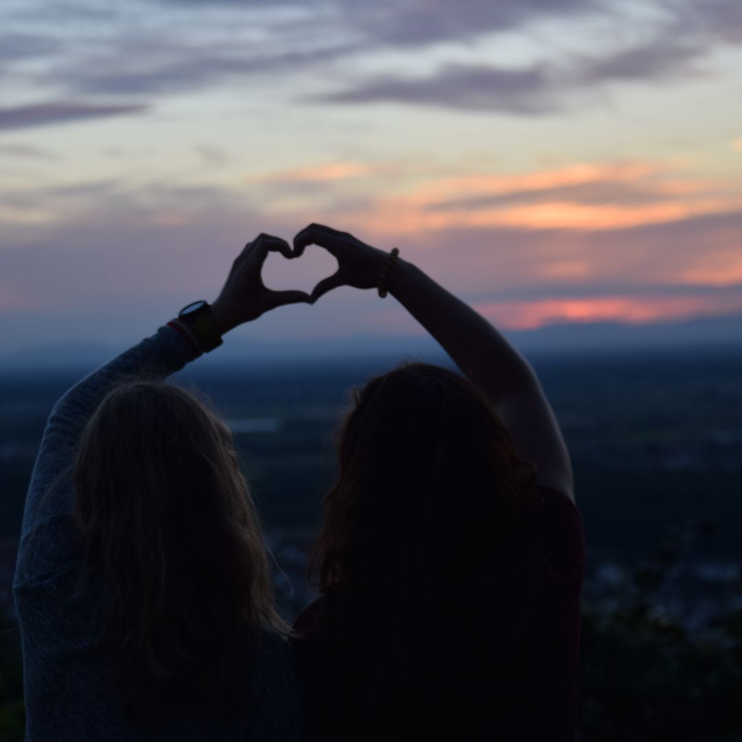 A silhouette of Two people making the shape of a heart with their hands in front of a warm evening sunset.