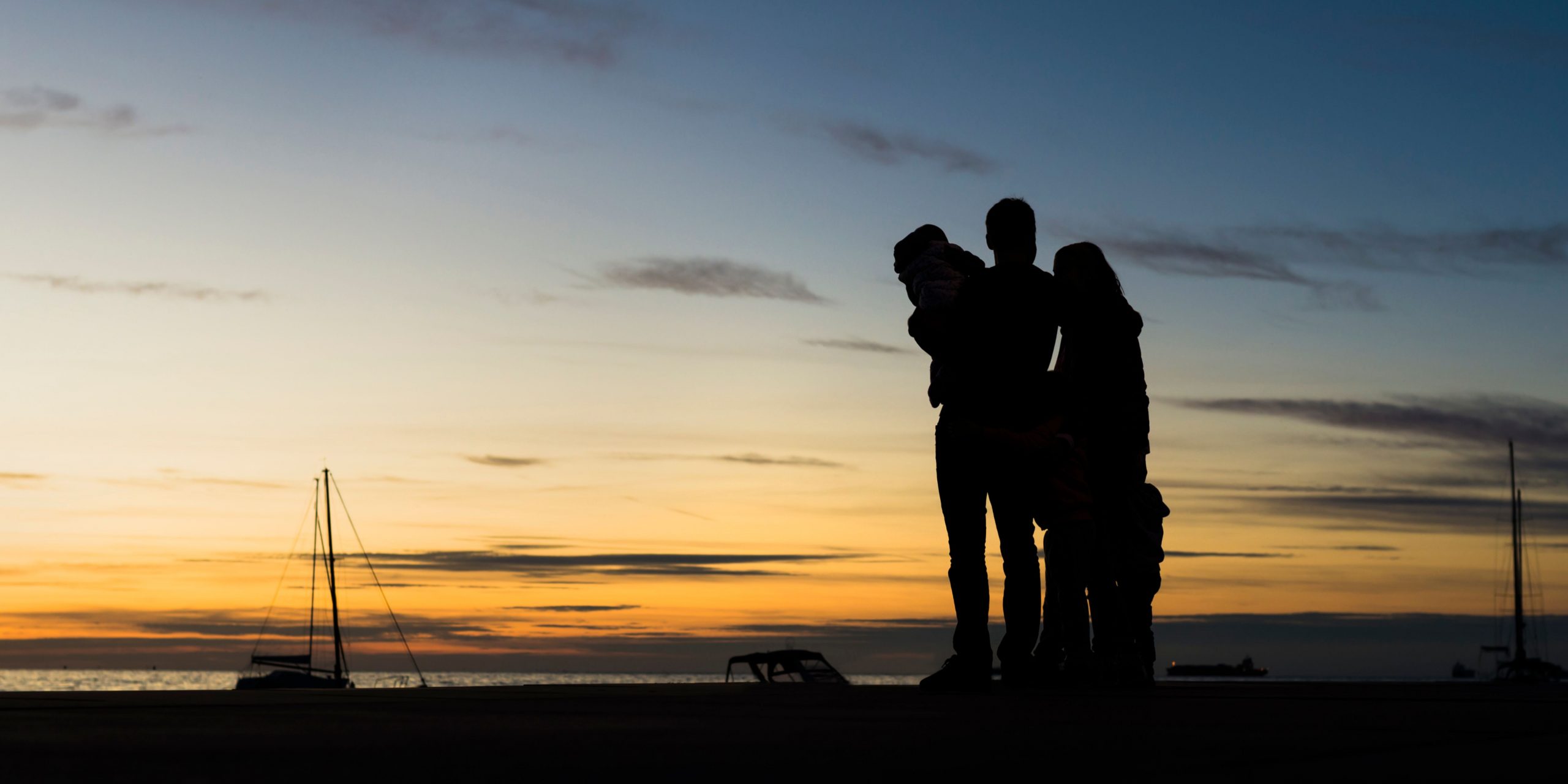 Two adults, one holding a child, silhouetted against a yellow and blue sunset by the sea.