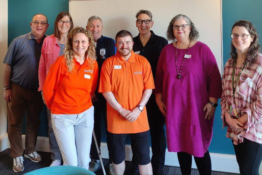 A group of 8 people stand smiling, two wearing Grassroots Suicide Prevention shirts.