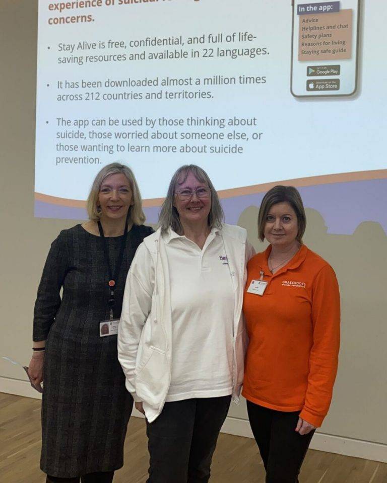 Three women stand smiling in front of a projection for the Stay Alive app.