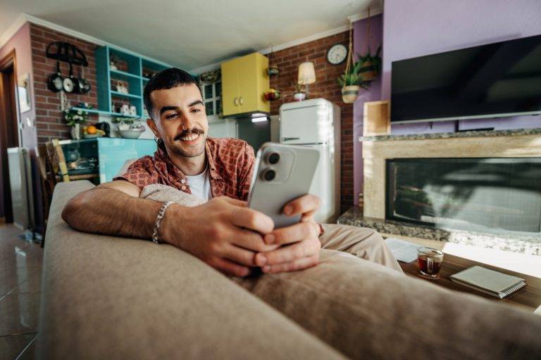 Cheerful hipster man using smart phone while relaxing on a sofa at home stock photo